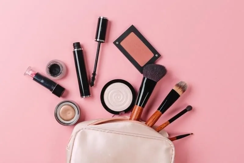 Make-uptasje met mascara, lippenstift, blush, highlighter en kwasten op een roze achtergrond