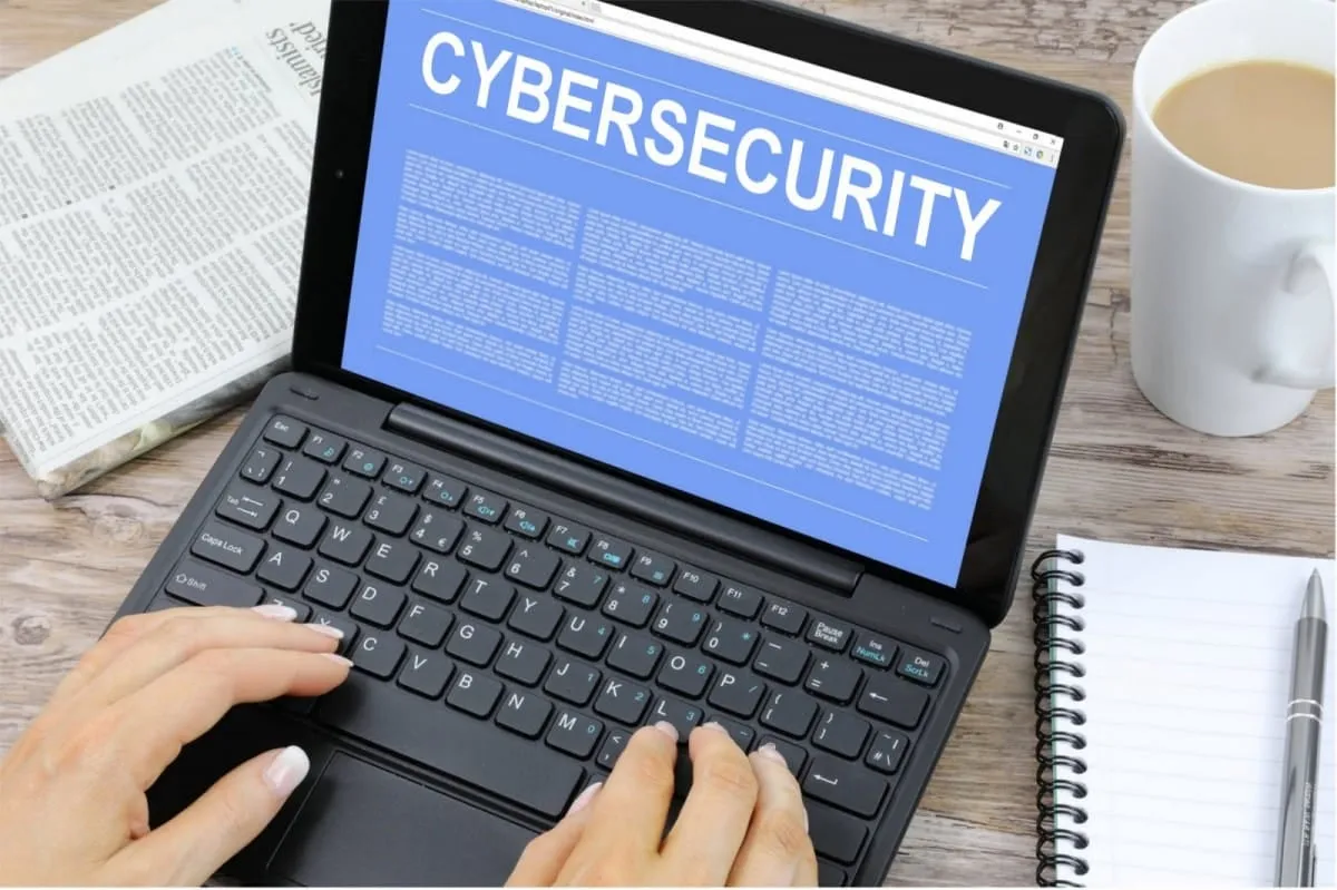 Handen typen op een laptop met het woord cybersecurity op het scherm, naast een kop koffie en een notitieblok