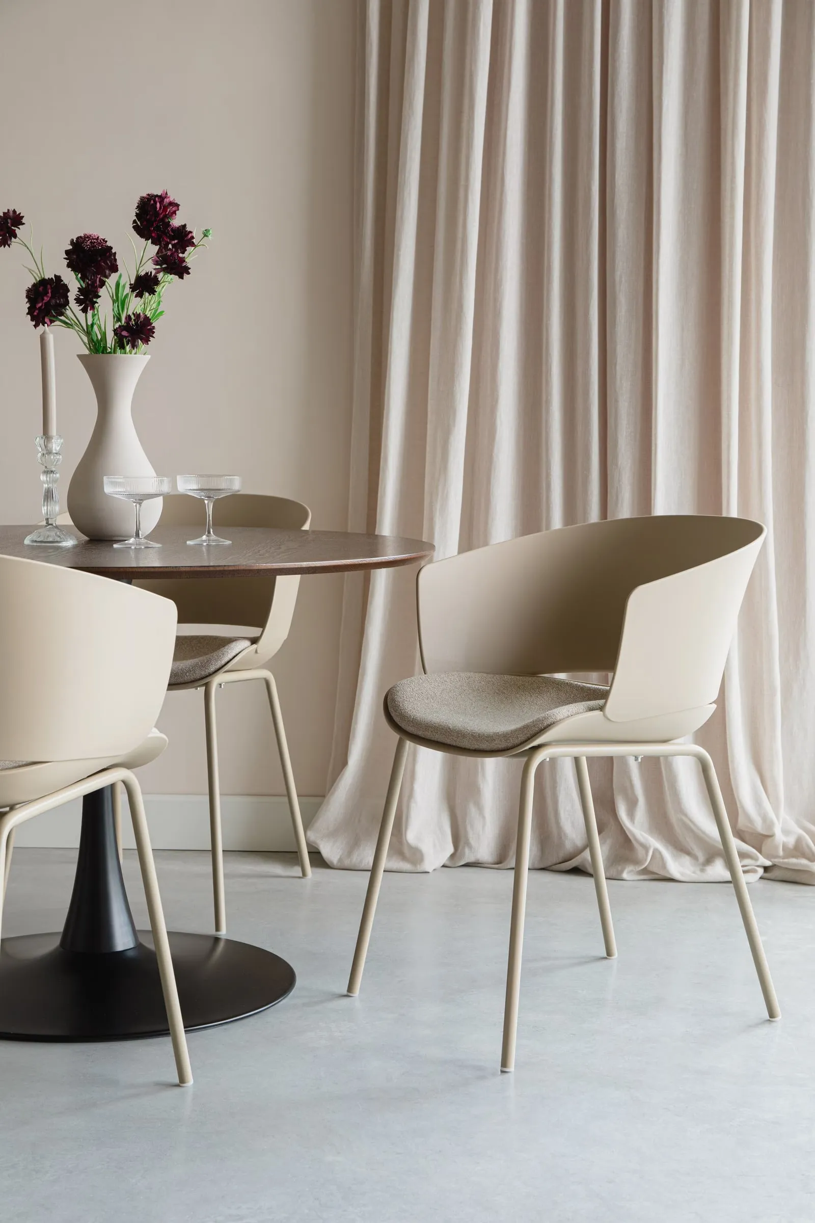Twee beige Japandi eetkamerstoelen met slanke poten rond een donkere ronde eettafel bij een linnen gordijn